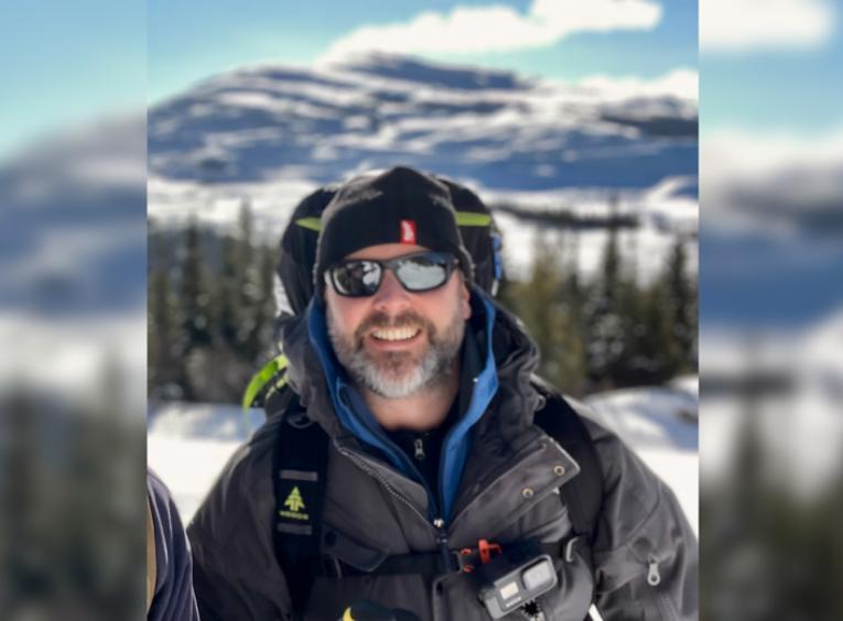 Man in sunglasses and winter gear stands in front of mountain backdrop